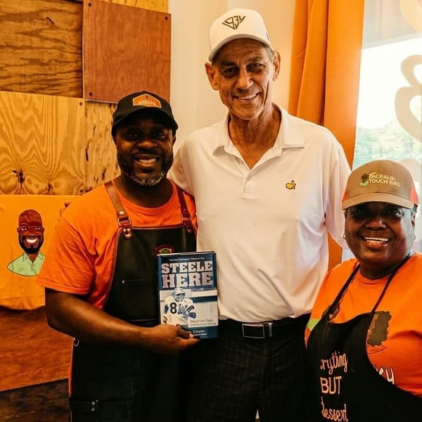 Three smiling people, two in orange shirts and aprons, pose with a man holding a book.