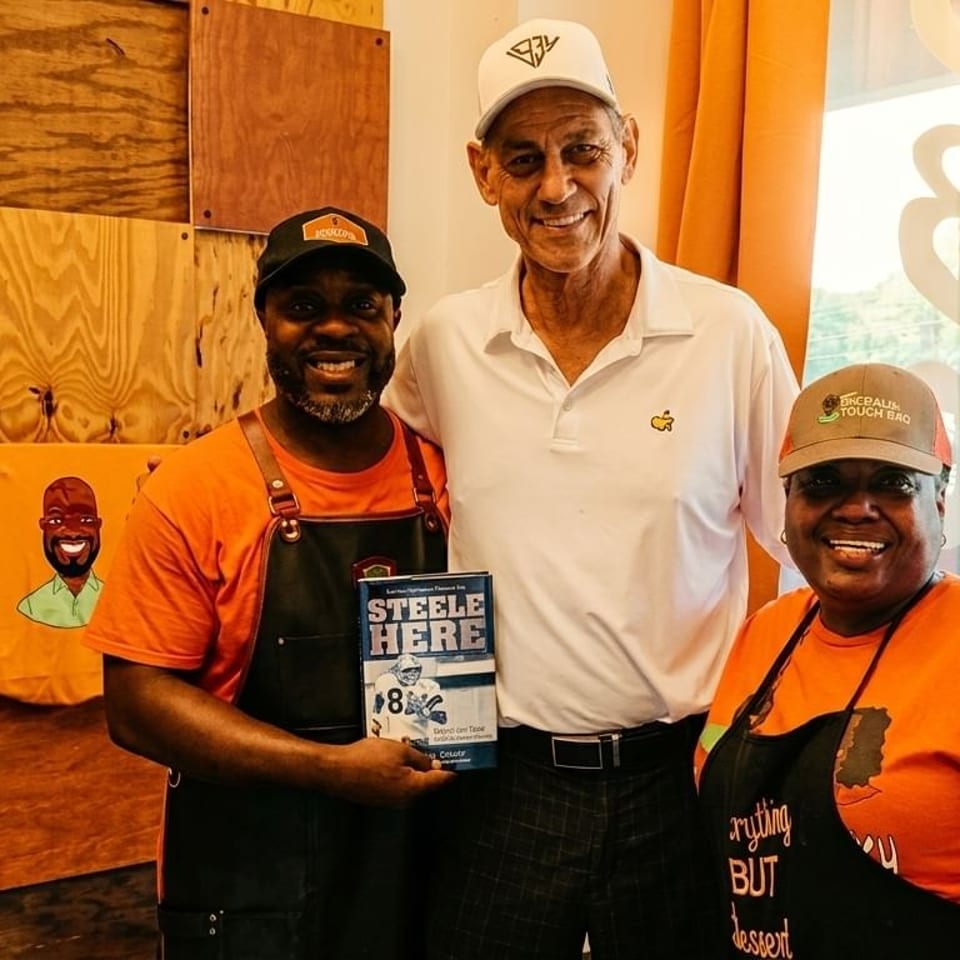 Three smiling people, two in orange shirts and aprons, pose with a man holding a book.