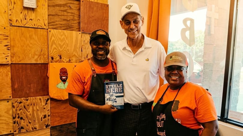 Three smiling people, two in orange shirts and aprons, pose with a man holding a book.