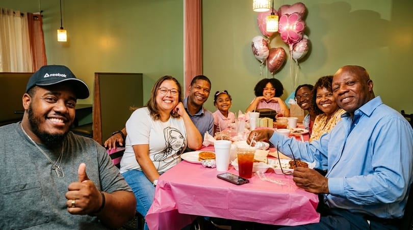 A large family smiles at the camera while sitting around a table with food and drinks.