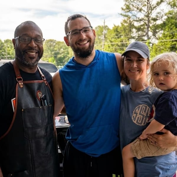 A family with a young child poses with a restaurant worker in an apron outside.