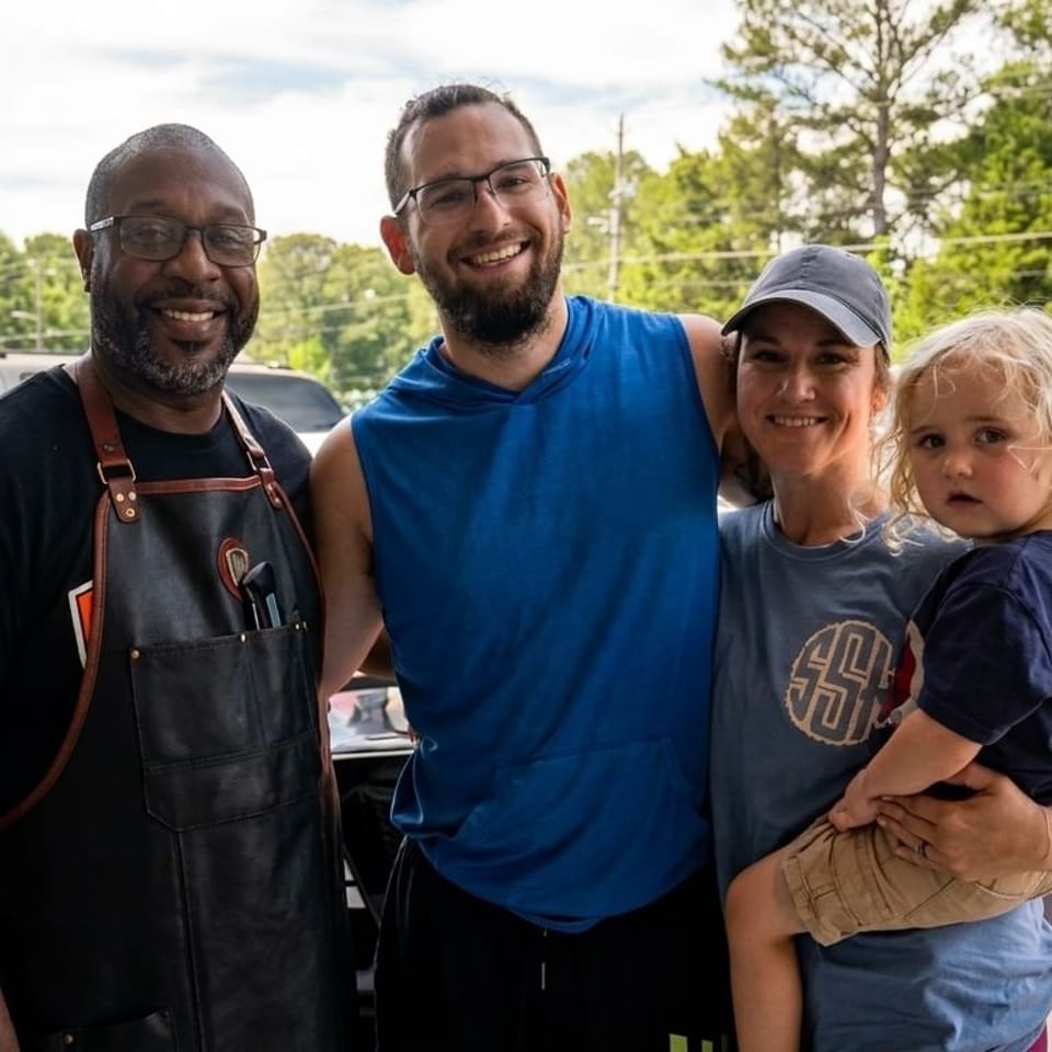 A family with a young child poses with a restaurant worker in an apron outside.