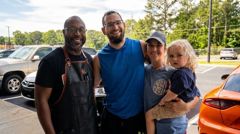 A family with a young child poses with a restaurant worker in an apron outside.