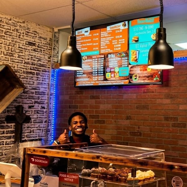 Smiling man giving thumbs up behind a counter with cupcakes, brick walls, and menu screens.