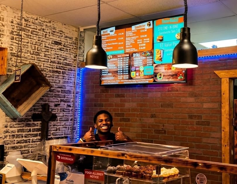 Smiling man giving thumbs up behind a counter with cupcakes, brick walls, and menu screens.