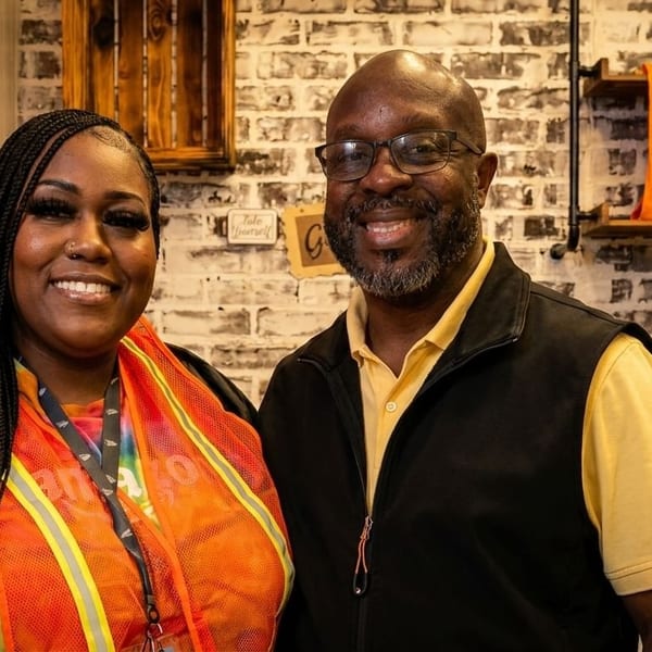 Two smiling people, a woman and a man, stand in front of a brick wall with shelves.