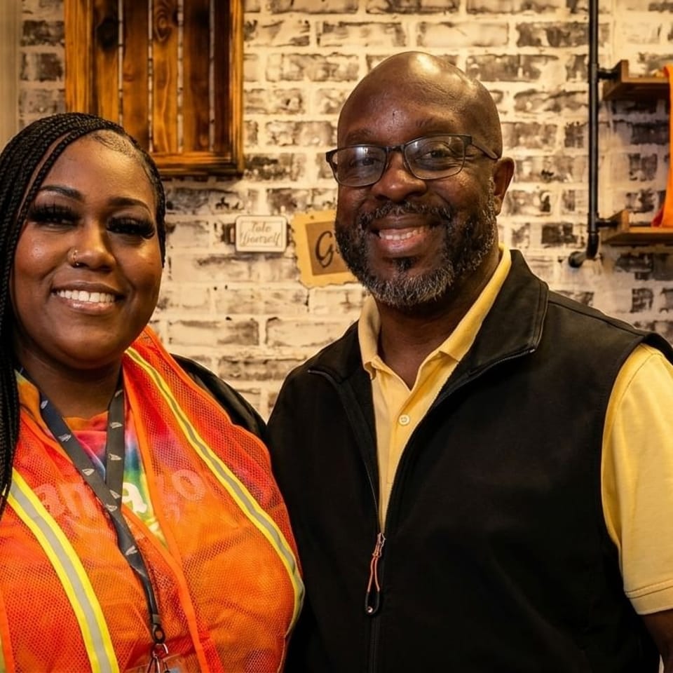 Two smiling people, a woman and a man, stand in front of a brick wall with shelves.