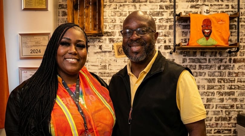 Two smiling people, a woman and a man, stand in front of a brick wall with shelves.