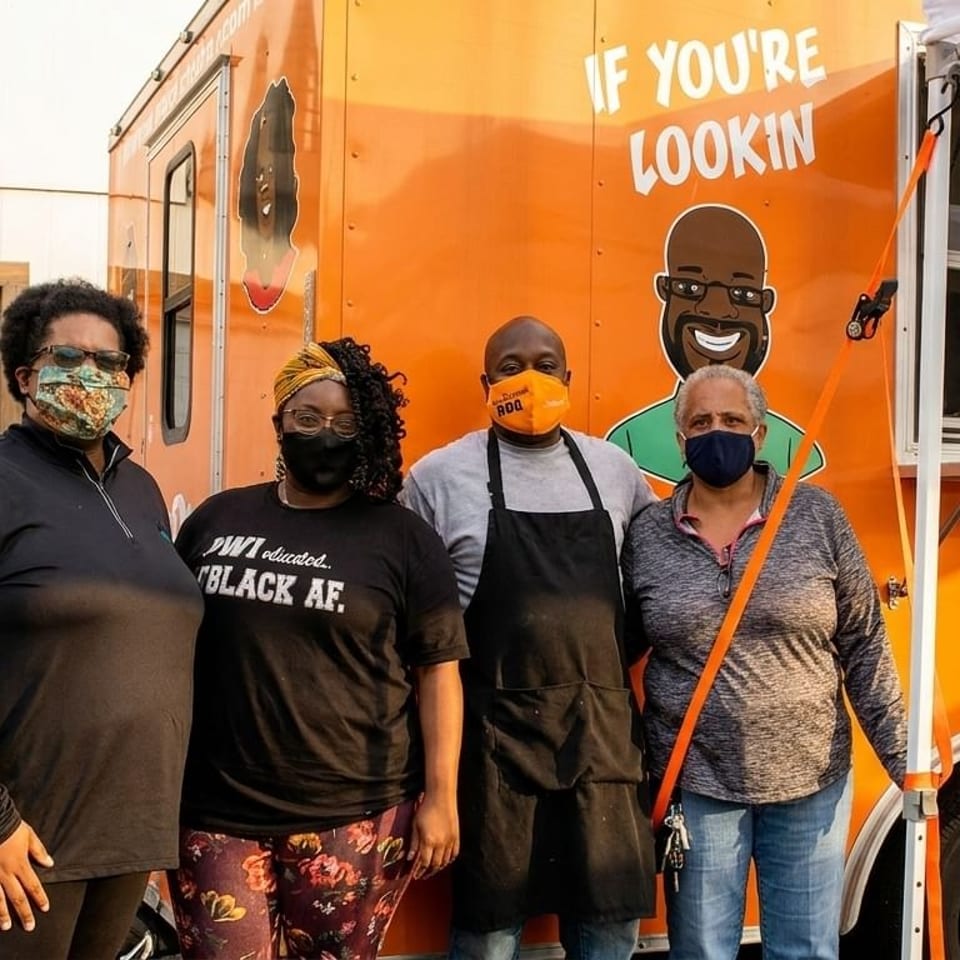 Four people wearing masks stand in front of an orange food truck with cartoon characters.