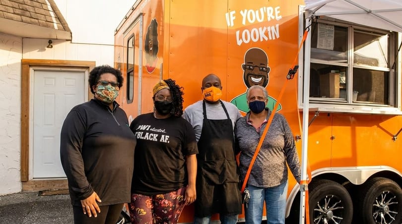 Four people wearing masks stand in front of an orange food truck with cartoon characters.