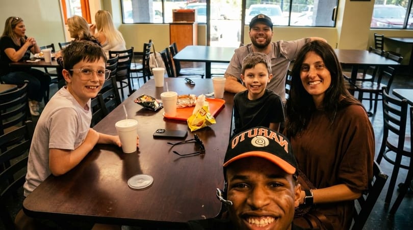 A family and friends smiling at a table in a casual restaurant, with food and drinks.