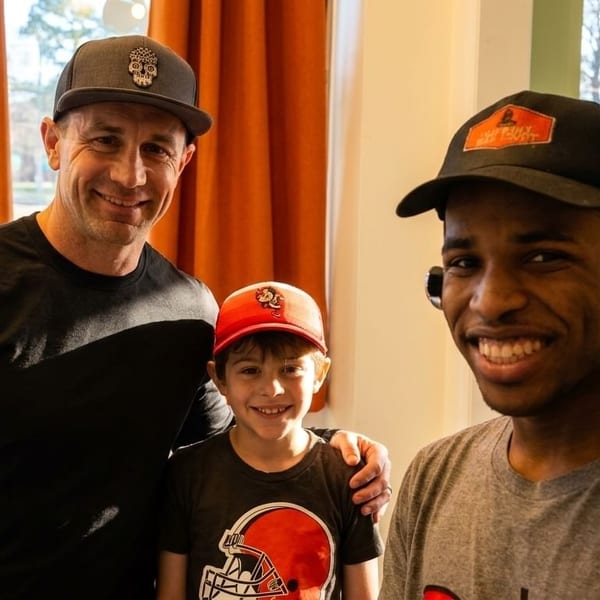 Three smiling males, two adults and one child, pose together indoors, wearing hats and casual shirts.