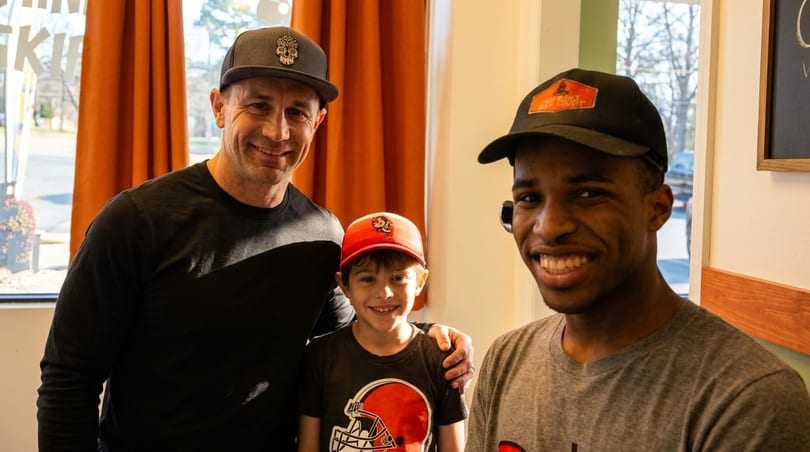 Three smiling males, two adults and one child, pose together indoors, wearing hats and casual shirts.