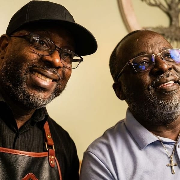 Two smiling Black men, one in a black cap and apron, the other in a light blue polo.