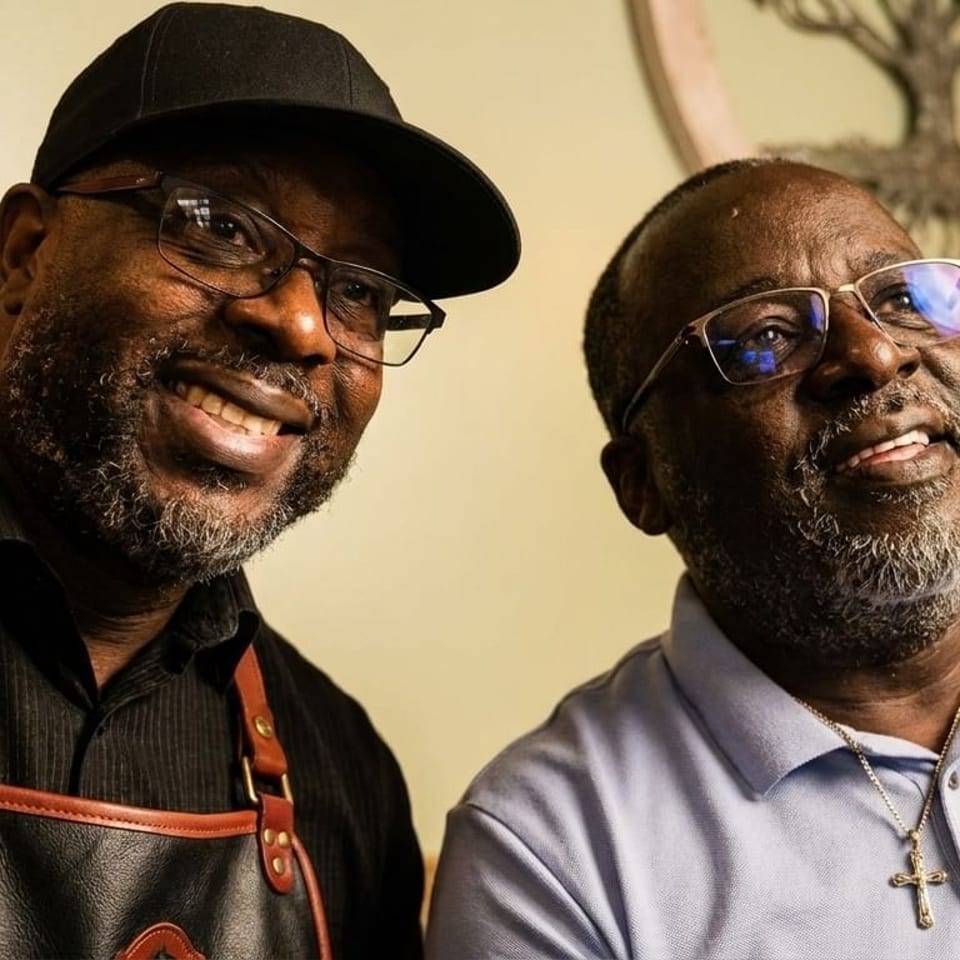 Two smiling Black men, one in a black cap and apron, the other in a light blue polo.