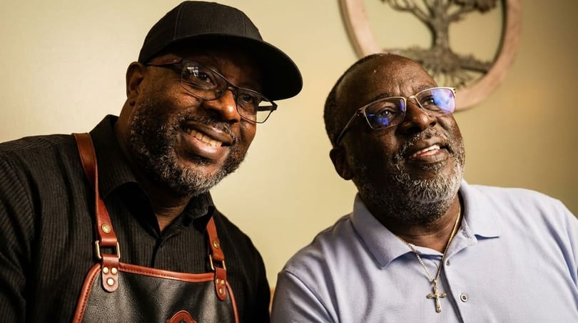 Two smiling Black men, one in a black cap and apron, the other in a light blue polo.