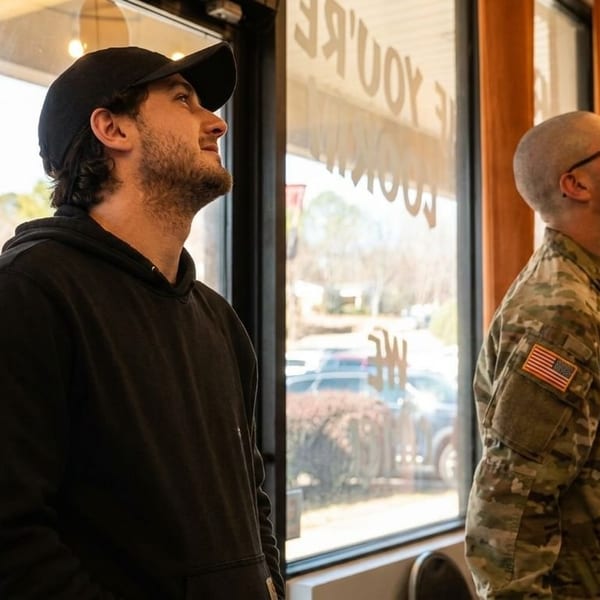 Customers, including a man in military uniform, look up inside a restaurant with large windows.