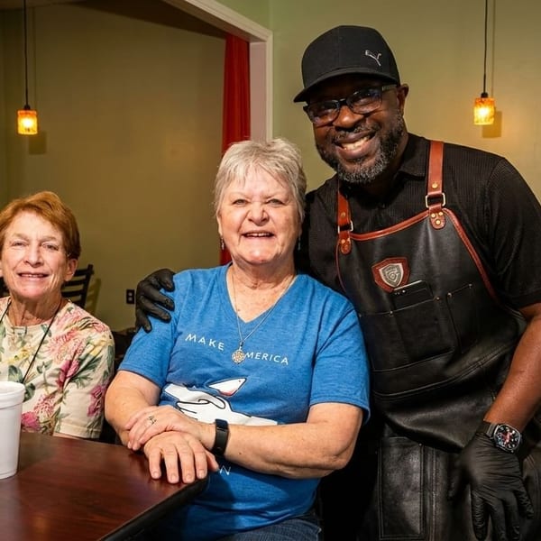 A smiling chef in an apron poses with two happy female customers at a restaurant table.
