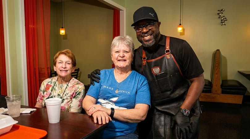 A smiling chef in an apron poses with two happy female customers at a restaurant table.