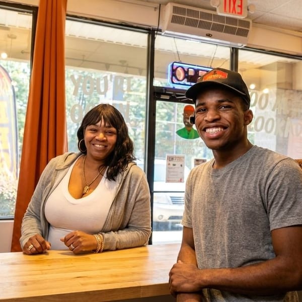Two smiling people, a woman and a man, stand behind a wooden counter in a restaurant.