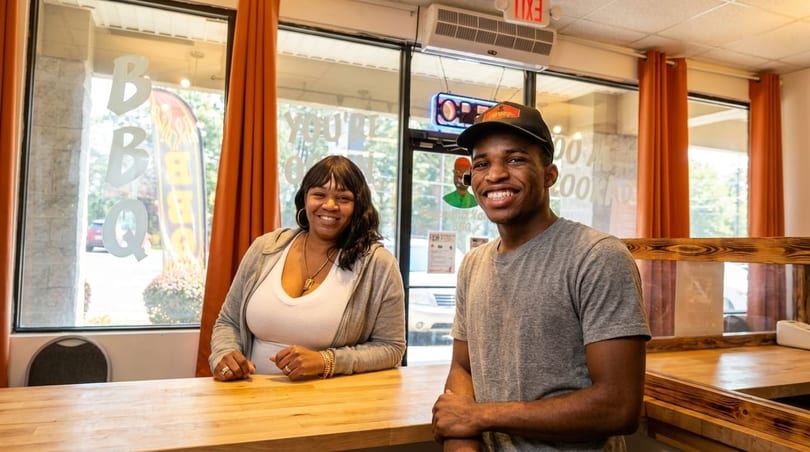 Two smiling people, a woman and a man, stand behind a wooden counter in a restaurant.