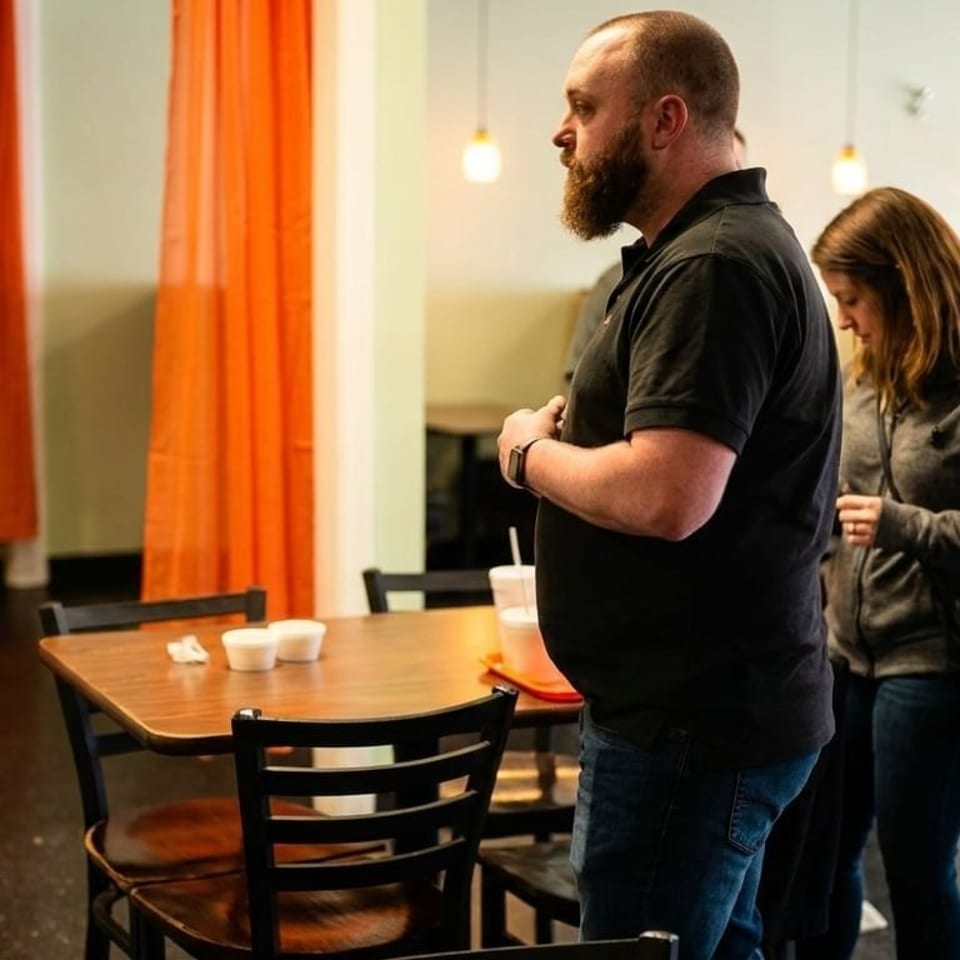 Two customers stand in a restaurant dining room with orange curtains and wooden tables.