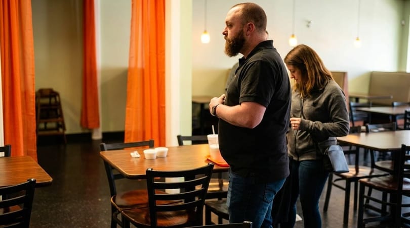 Two customers stand in a restaurant dining room with orange curtains and wooden tables.