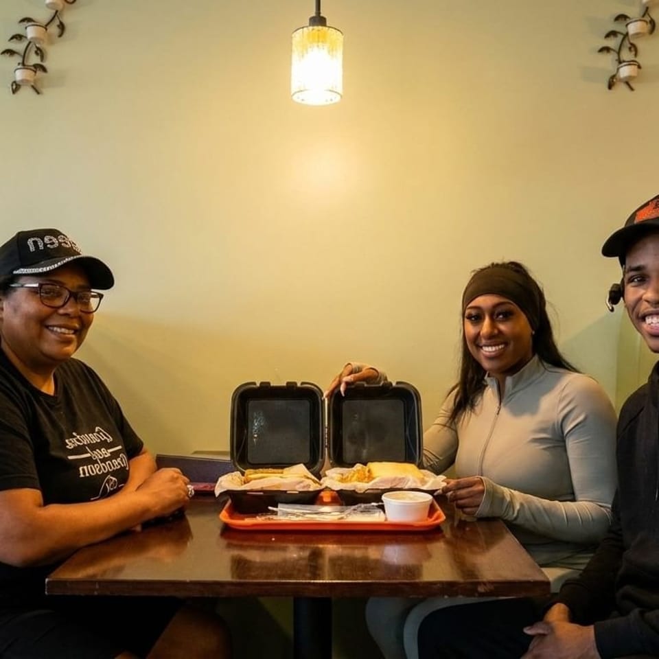 Three smiling people sit at a booth table with open takeout containers of food.