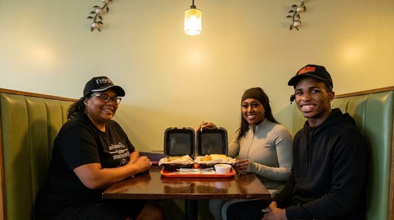 Three smiling people sit at a booth table with open takeout containers of food.