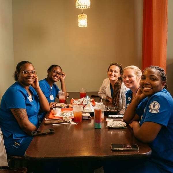 Five smiling women in scrubs and casual clothes sit around a restaurant table with drinks and food.