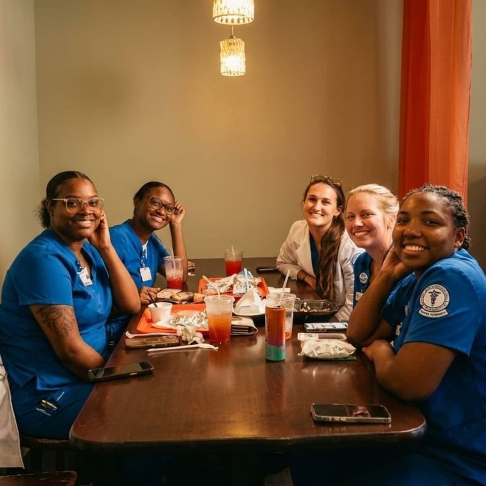 Five smiling women in scrubs and casual clothes sit around a restaurant table with drinks and food.