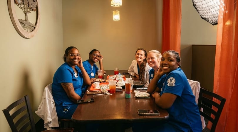 Five smiling women in scrubs and casual clothes sit around a restaurant table with drinks and food.