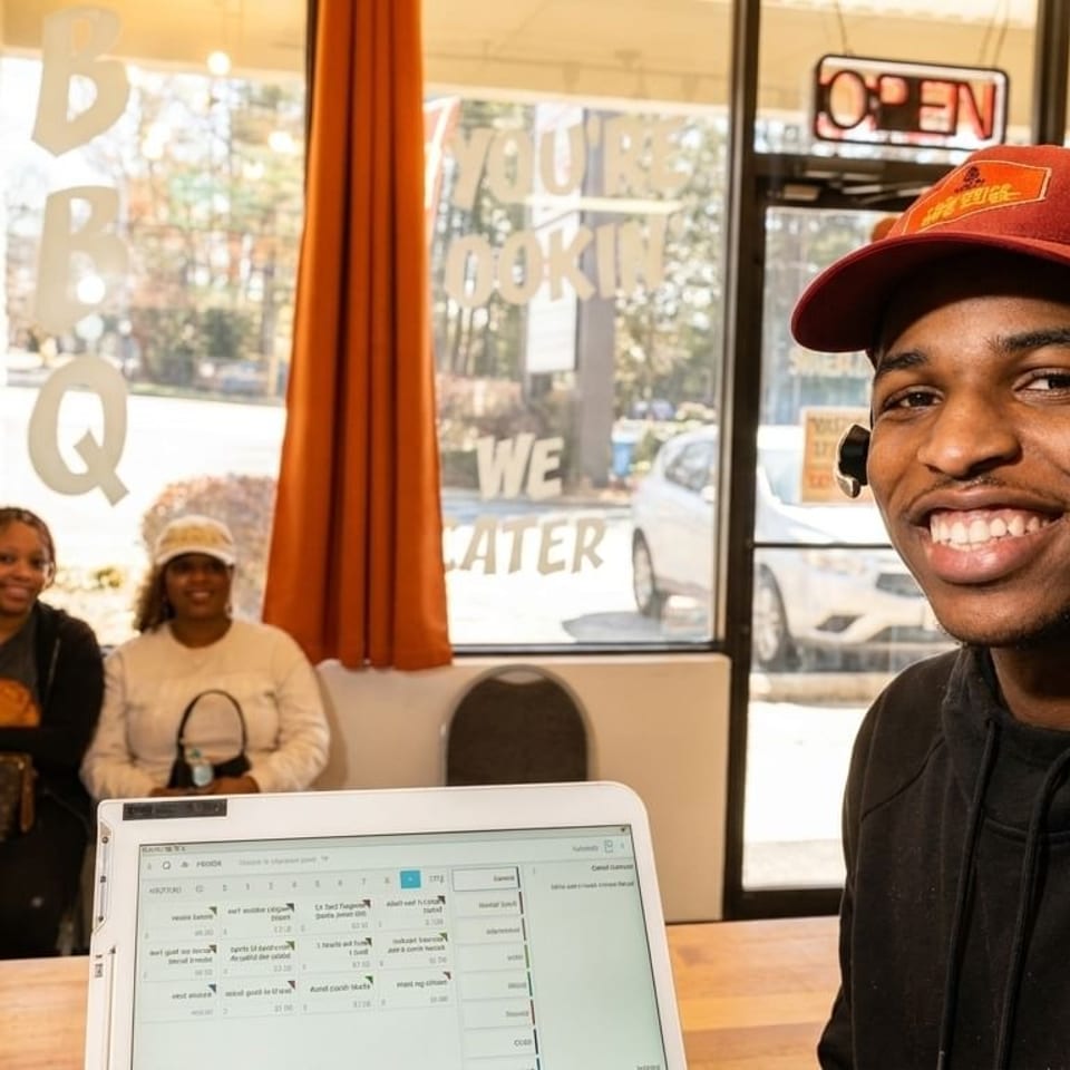 Smiling man in a red hat at a counter, with two women seated behind him in a restaurant.