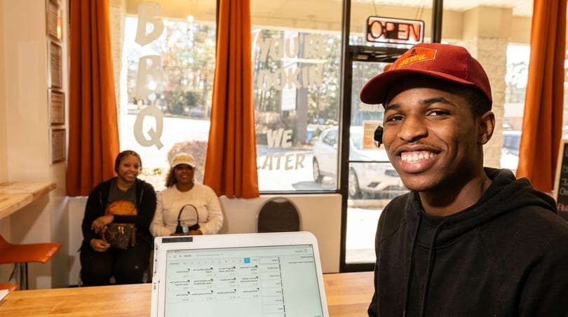 Smiling man in a red hat at a counter, with two women seated behind him in a restaurant.