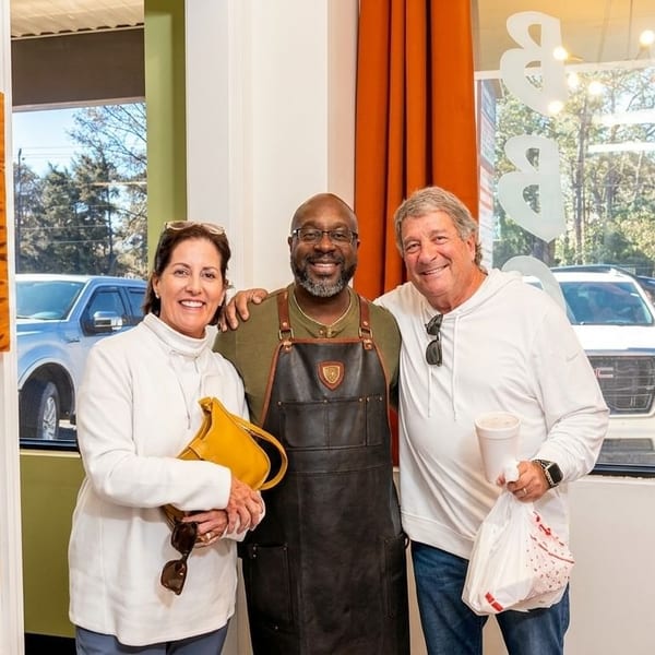 A chef in a leather apron stands between two smiling customers in a restaurant.