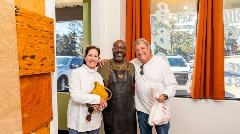 A chef in a leather apron stands between two smiling customers in a restaurant.