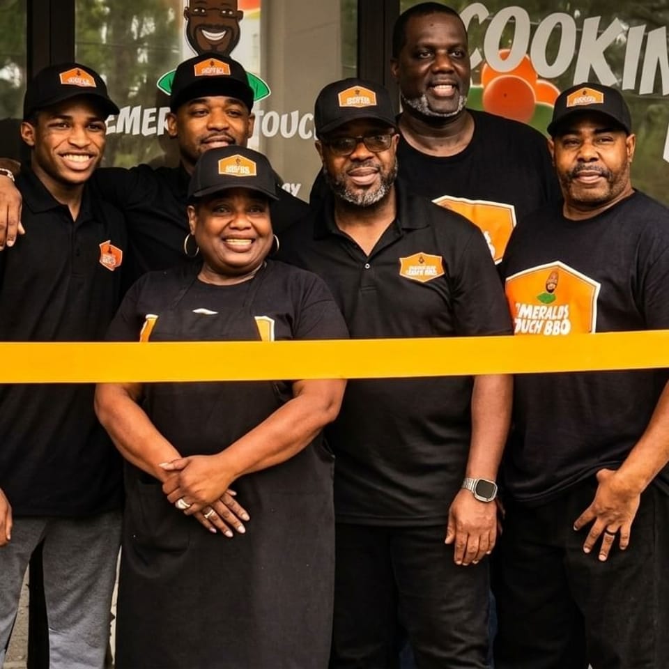 Six smiling people in black shirts and hats stand behind a yellow ribbon in front of a restaurant.