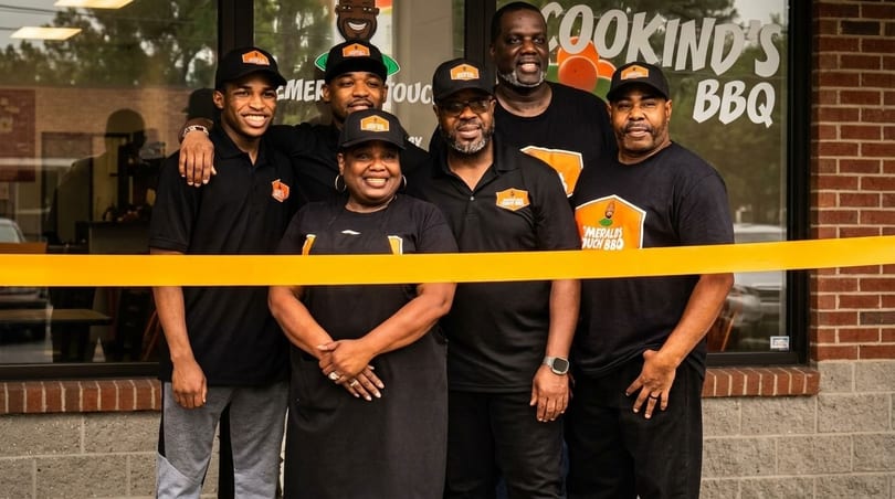 Six smiling people in black shirts and hats stand behind a yellow ribbon in front of a restaurant.