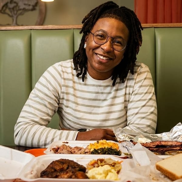 Smiling person with dreadlocks and glasses sitting at a booth with takeout containers of barbecue.