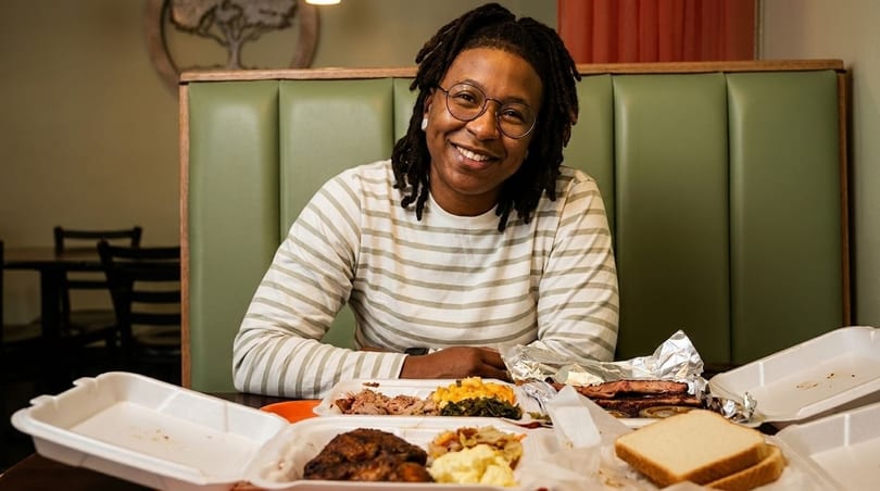Smiling person with dreadlocks and glasses sitting at a booth with takeout containers of barbecue.