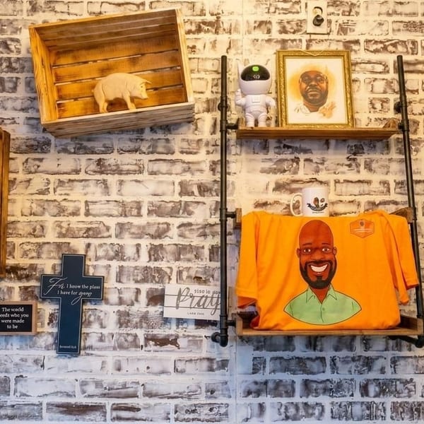 Decorated brick wall with wooden shelves displaying a pig figurine, framed portrait, robot, and t-shirt.