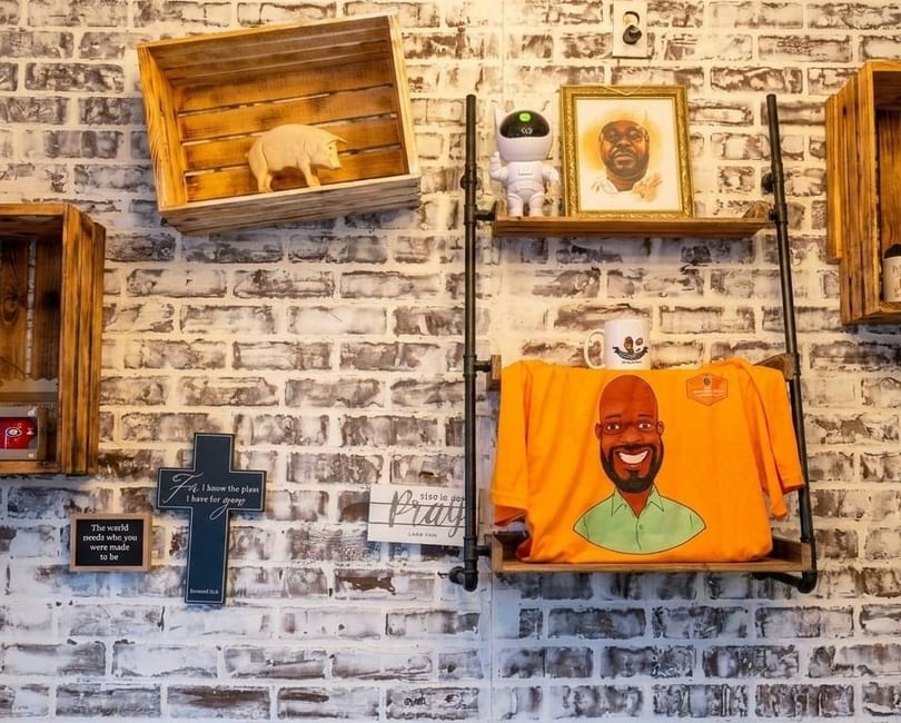 Decorated brick wall with wooden shelves displaying a pig figurine, framed portrait, robot, and t-shirt.