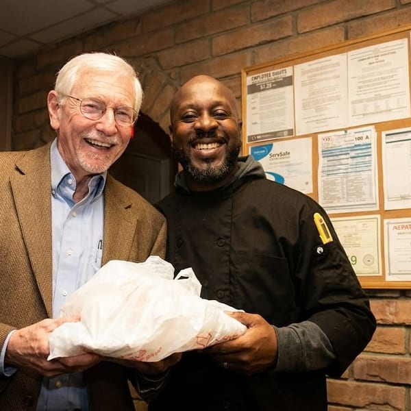 Two smiling men, one in a suit and one in a chef's coat, holding a white bag.