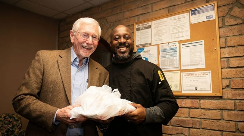 Two smiling men, one in a suit and one in a chef's coat, holding a white bag.