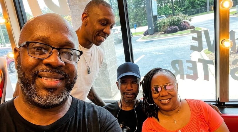 Four smiling people, two men and two women, pose for a selfie inside a restaurant with window views.