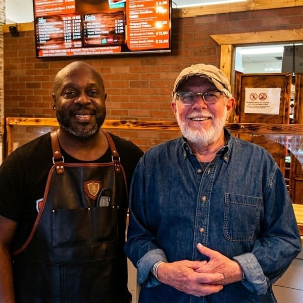 Two smiling men, one in an apron, stand behind a counter in a casual restaurant.
