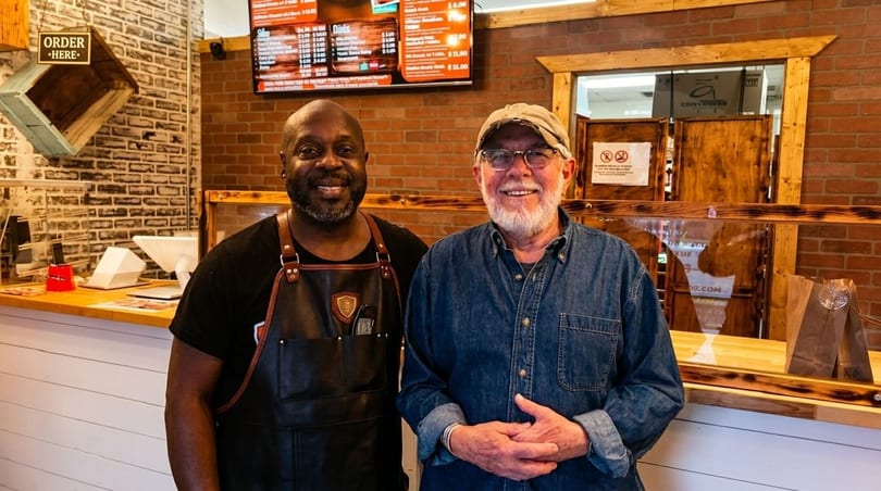 Two smiling men, one in an apron, stand behind a counter in a casual restaurant.