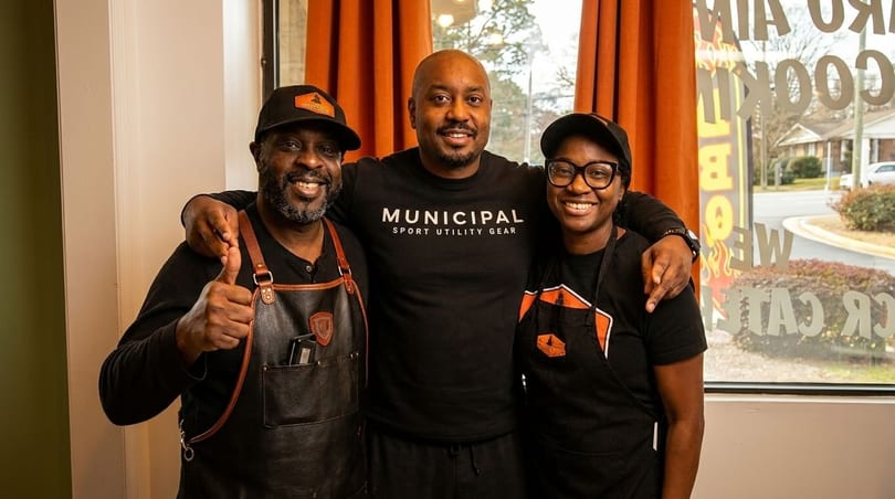 Three smiling restaurant staff members, two men and one woman, pose together in front of a window.